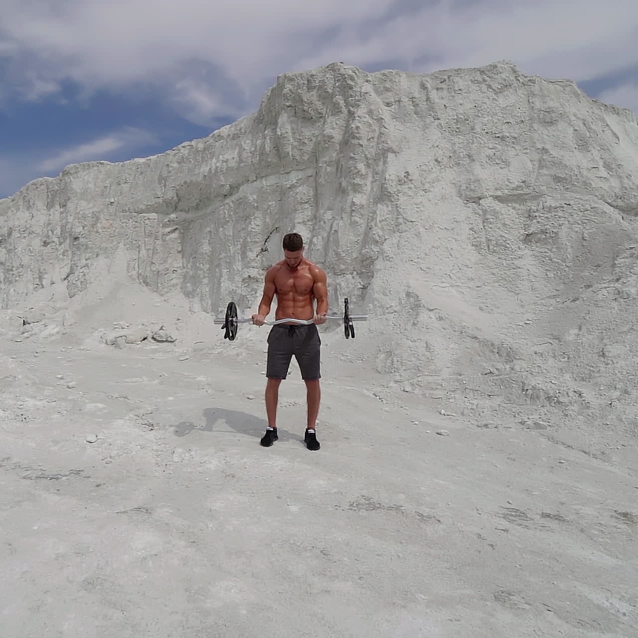Athletic guy with muscular body on mountain background outdoors. Beautiful view of white mountain and a strong man is lifting barbell under blue sky.
