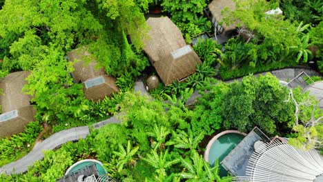 Birds-eye-view-of-luxurious-resort-over-hill-with-straw-roofs-surrounded-by-greenery-with-small-huts-and-houses-spread-across-the-city-with-calm-sea-and-mountains