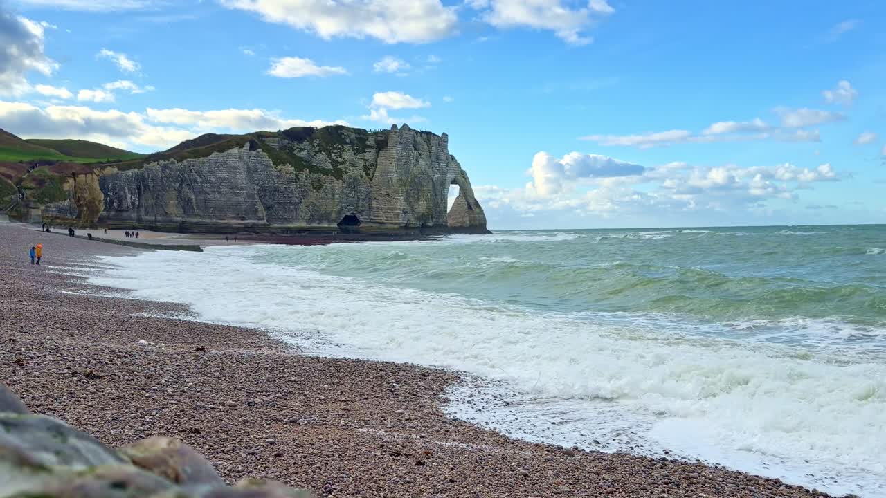 Static shot of ocean waves on the pebble beach with the Porte d'Aval cliff and natural arch in Étretat, Normandy, France