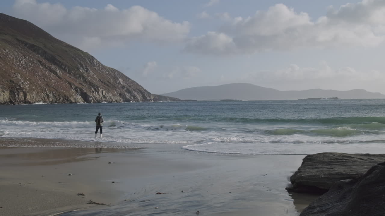 Super wide shot of a man standing on a beach in the water. Beautiful day with clouds and sun from the back. A few hills in the background.