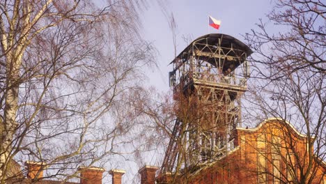 old metal mining tower with the czech flag on top, surrounded by bare trees against a clear sky