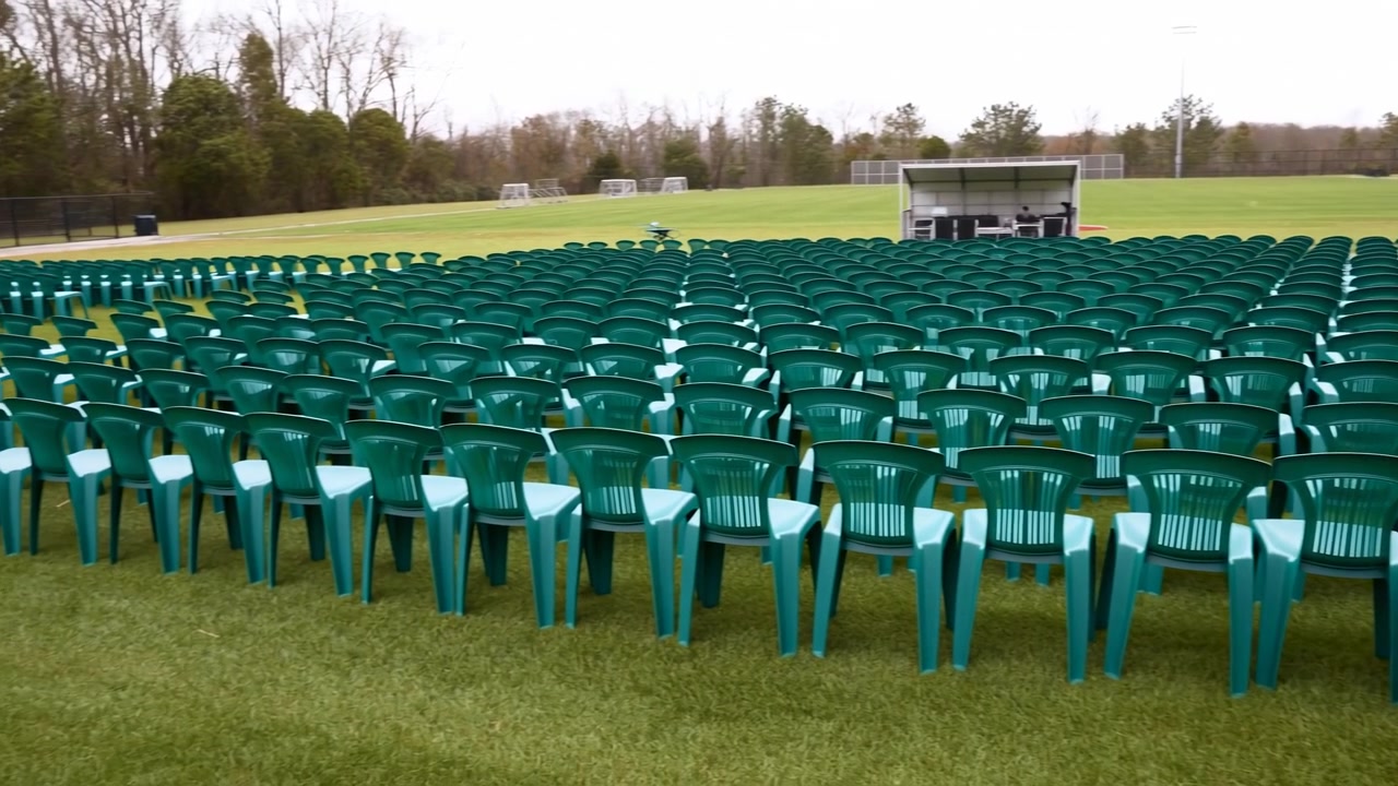 Hundreds of identical teal plastic chairs arranged in tight theater style rows across grassy sports field, facing small open sided stage or control booth with overcast light, creating orderly