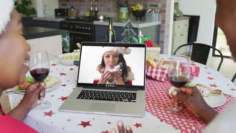 una pareja afroamericana con vino usando una computadora portátil para una videollamada de navidad con una mujer feliz en la pantalla