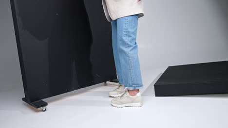 unrecognized female wearing jeans and jacket holding a bag. model in sports shoes standing near the black screen.