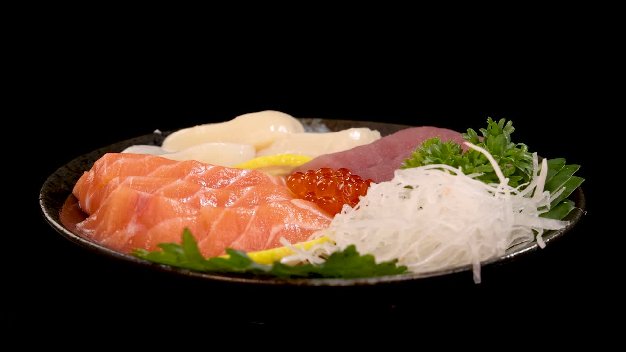 A plate of assorted sashimi and sushi, including salmon, tuna, scallop, and roe, rotates smoothly under studio lighting against a black background
