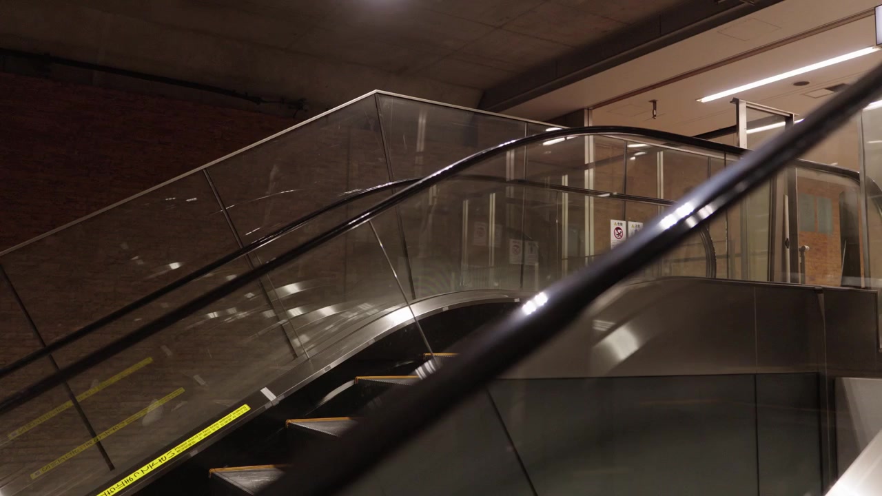 Escada rolante vazia em movimento com painéis de vidro e faixas de segurança amarelas dentro de uma moderna estação de metrô japonesa.