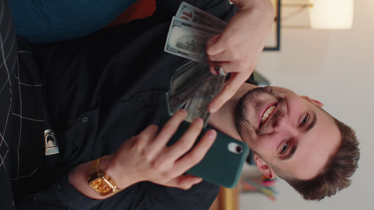 Giovane uomo barbuto sorridente e felice che conta denaro contante, usa lo smartphone, reddito, risparmi, vincita alla lotteria a casa