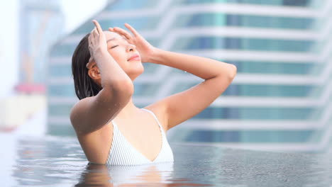 Bonita-Mujer-Asiática-En-Una-Piscina-Acariciando-Su-Cabello-Mojado-Con-Edificios-De-La-Ciudad-En-Segundo-Plano,-Cámara-Lenta