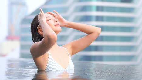 수영장에서 예쁜 아시아 여성이 도시 건물을 배경으로 젖은 머리를 애무하고 슬로우 모션