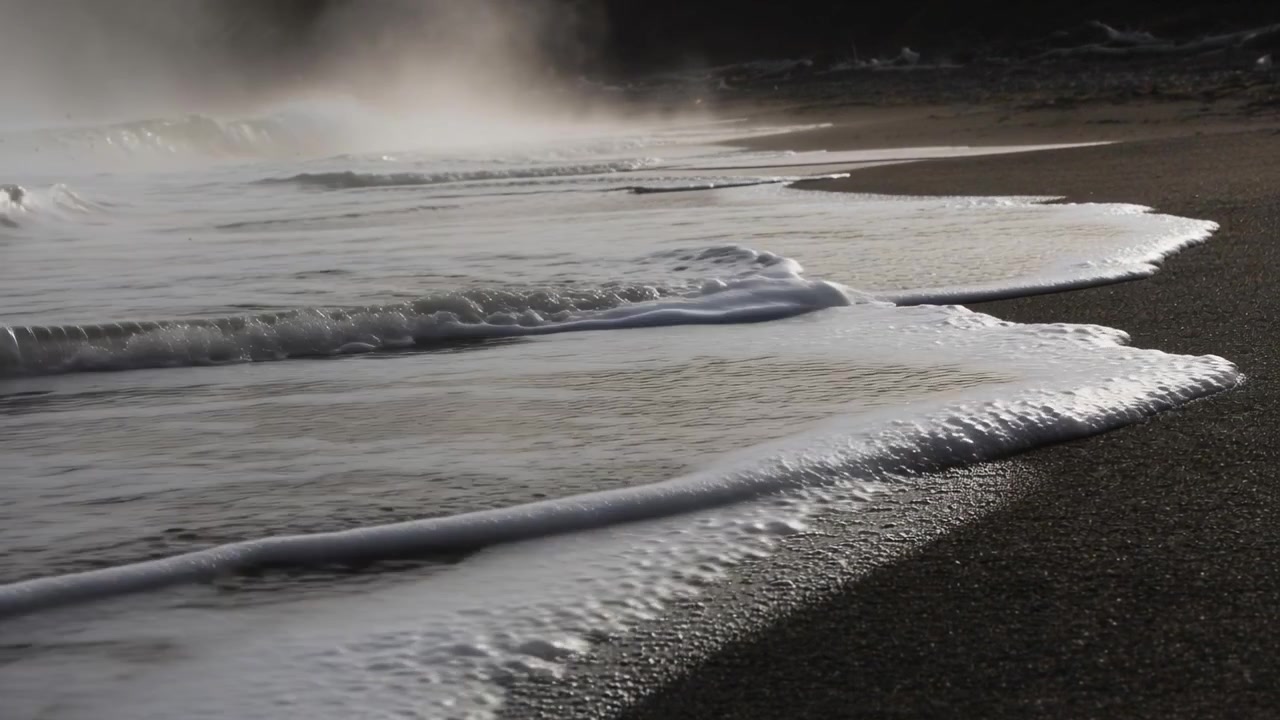 La spuma del mare che avanza si spinge su una spiaggia scura di ciottoli, con la mareggiata in arrivo che crea bolle e nebbia.