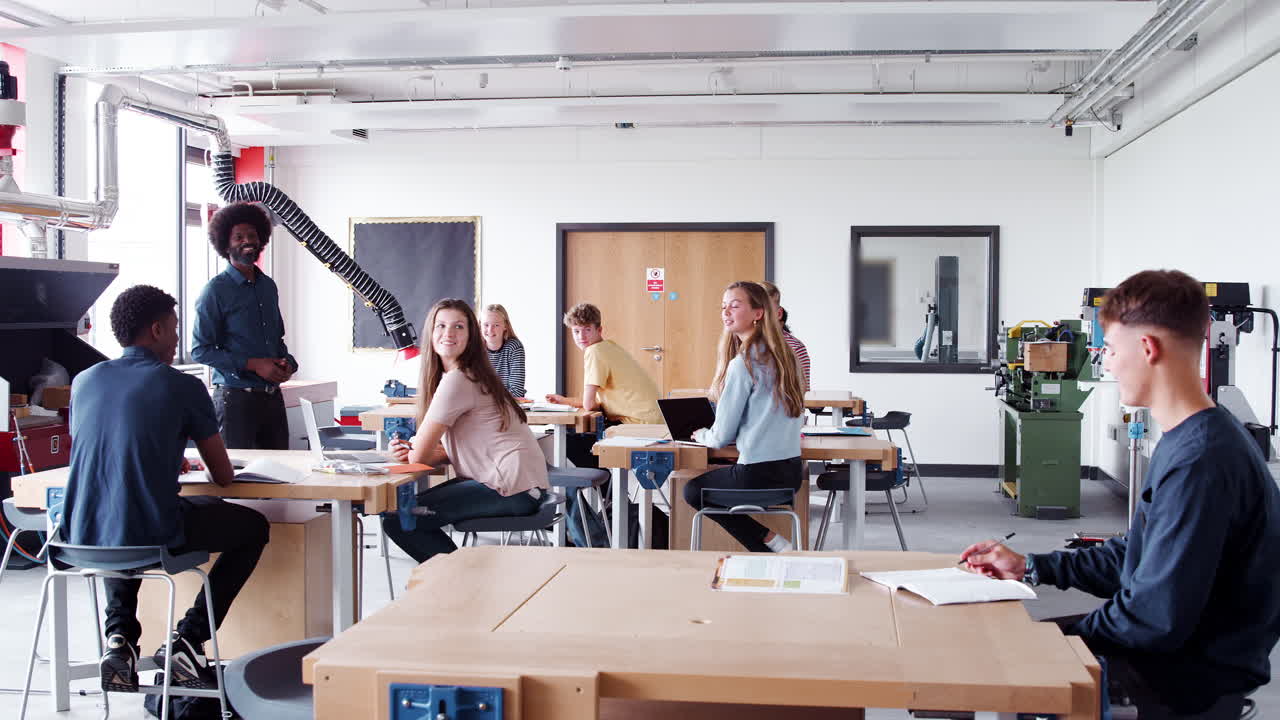 디자인 과 기술 수업 에서 작업 벤치 에 앉아 있는 고등학교 학생 들 의 그룹 과 이야기 하는 교사