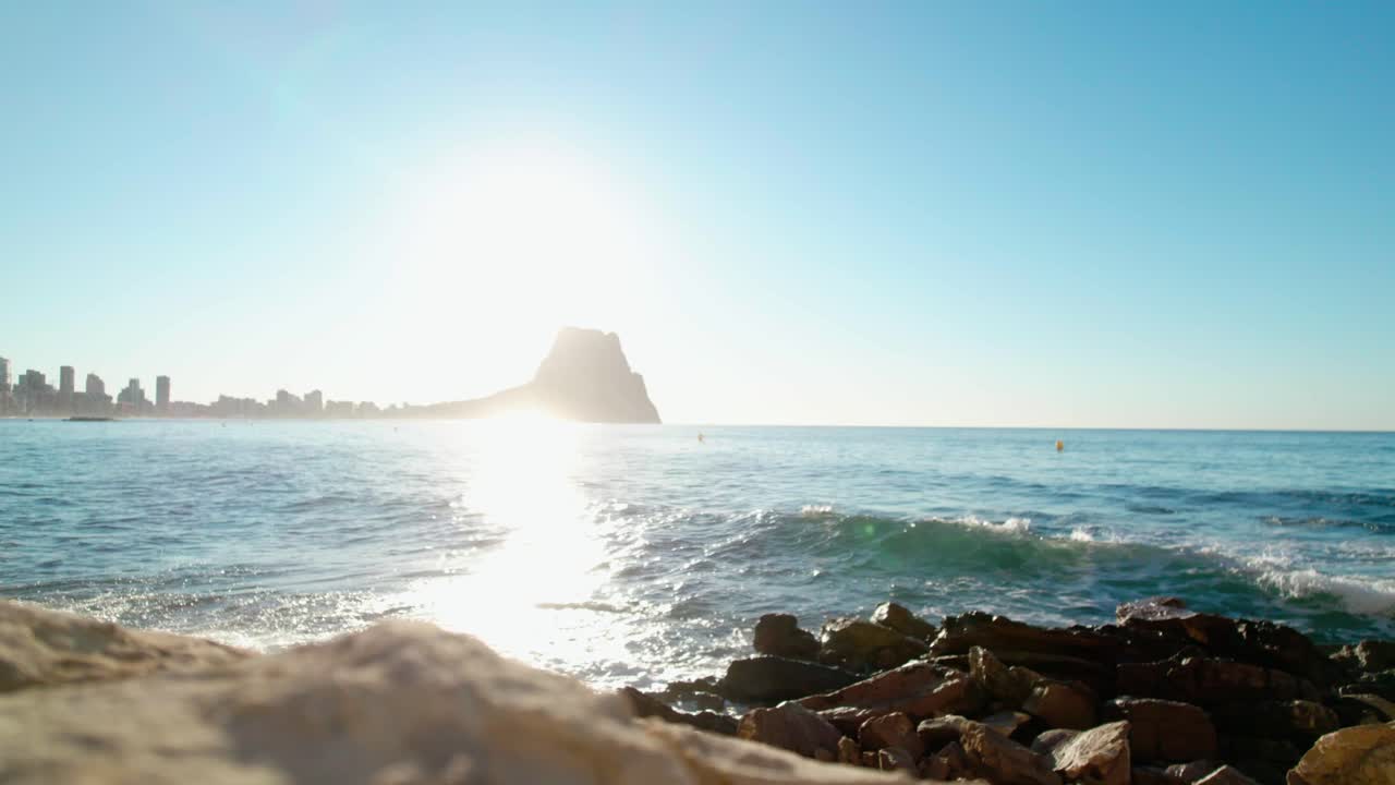 Waves Crashing On The Rocky Shores With Peñón de Ifach Rock In The Background In Calpe, Alicante, Spain. Static Shot