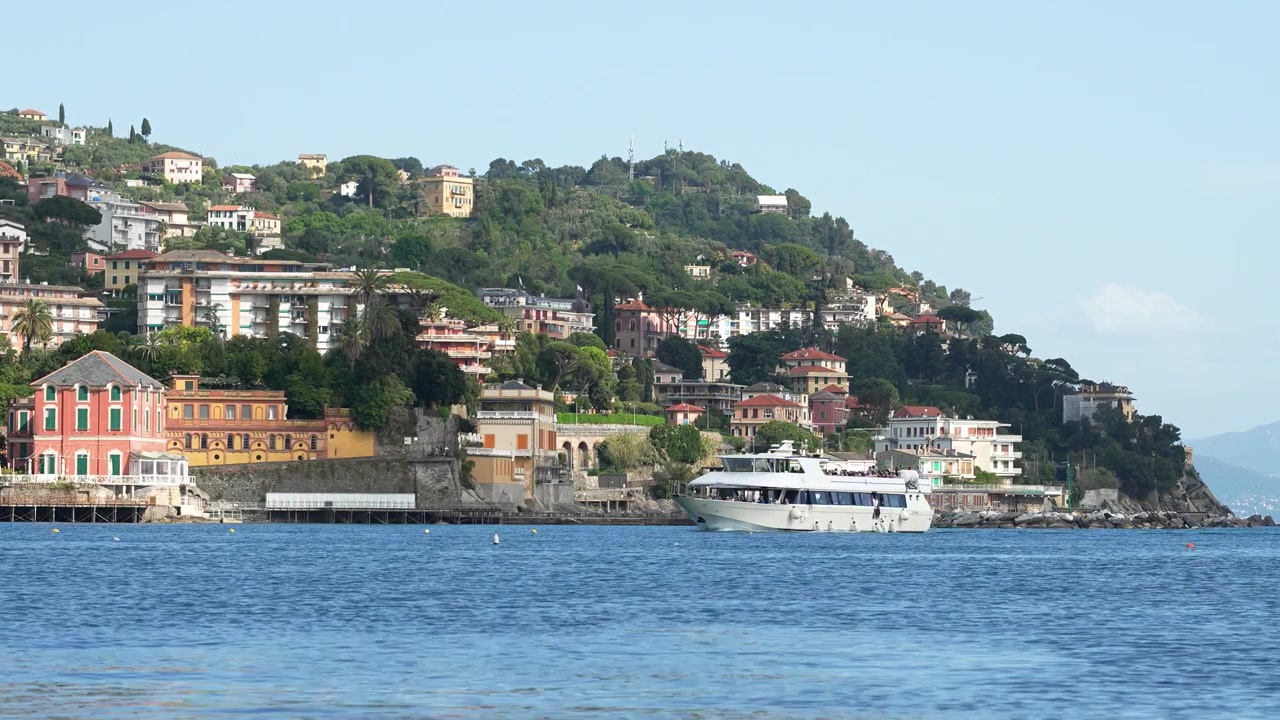 Riprese grandangolari della costa panoramica di Rapallo, Italia, con ville colorate sulle colline, vegetazione rigogliosa e un traghetto bianco che naviga su acque calme e azzurre sotto la luce del sole.
