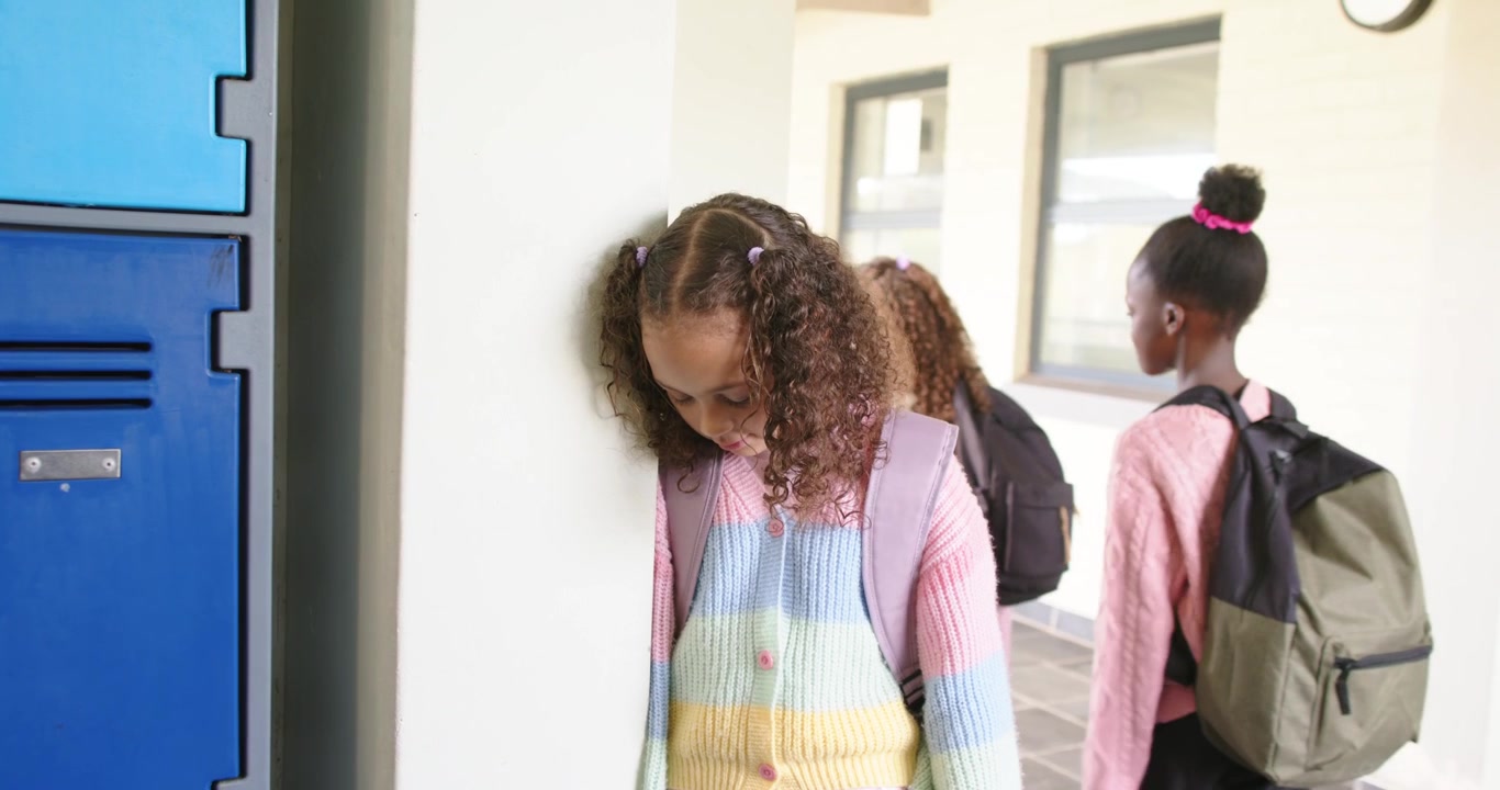 African American girls passing by, causing curly girl leaning on pillar by lockers, looking down
