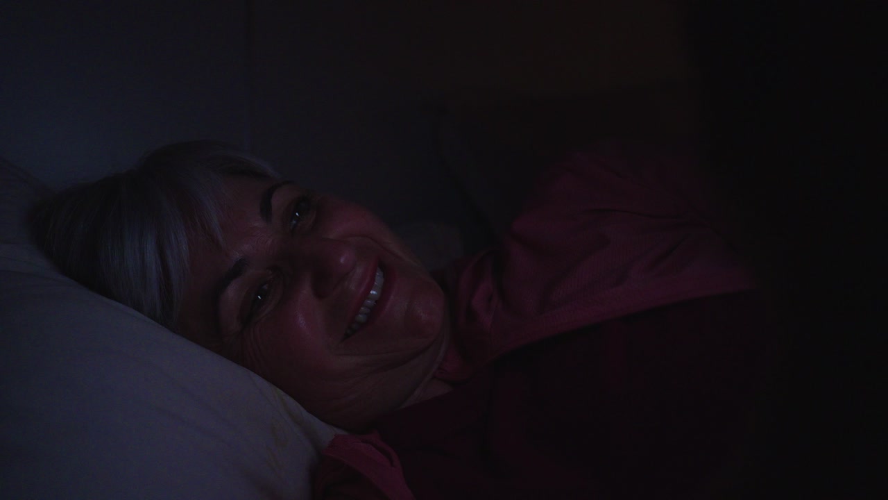 Mujer mayor feliz sonriendo mientras usa el teléfono en un dormitorio oscuro