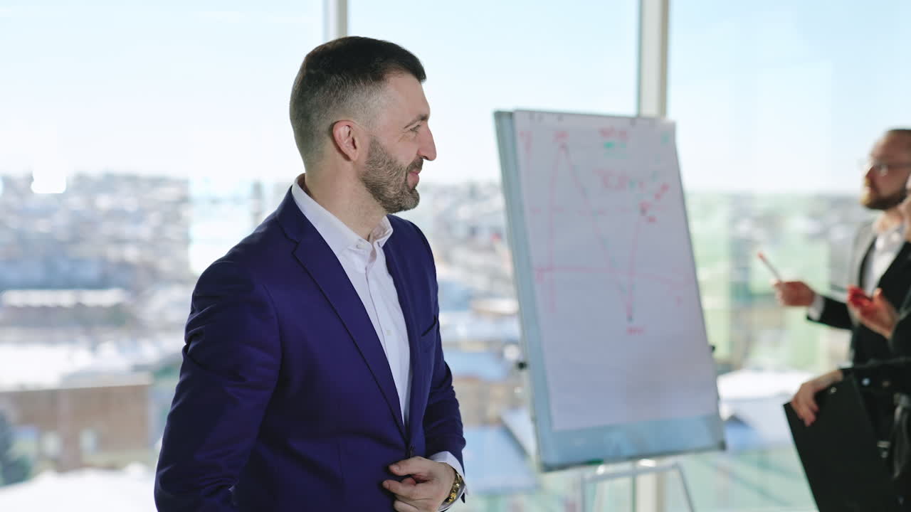Mid-aged bearded man in suit talks firstly to camera and then aside. Portrait of a confident male business person crossing hands on his chest. Office employees at backdrop in blur.
