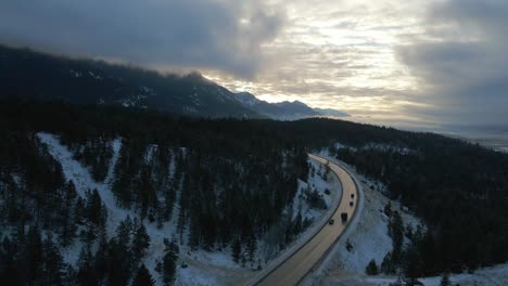 early morning sunrise aerial reveal of cars travelling along snow-covered mountain cariboo highway 95 with reflections on road in mystical, cloudy scene in british columbia, canada