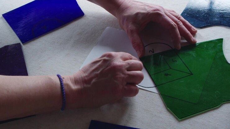 An art teacher shows her student how to trace a design onto a piece of green stained glass during an art class. A template of a heart is visible below.
