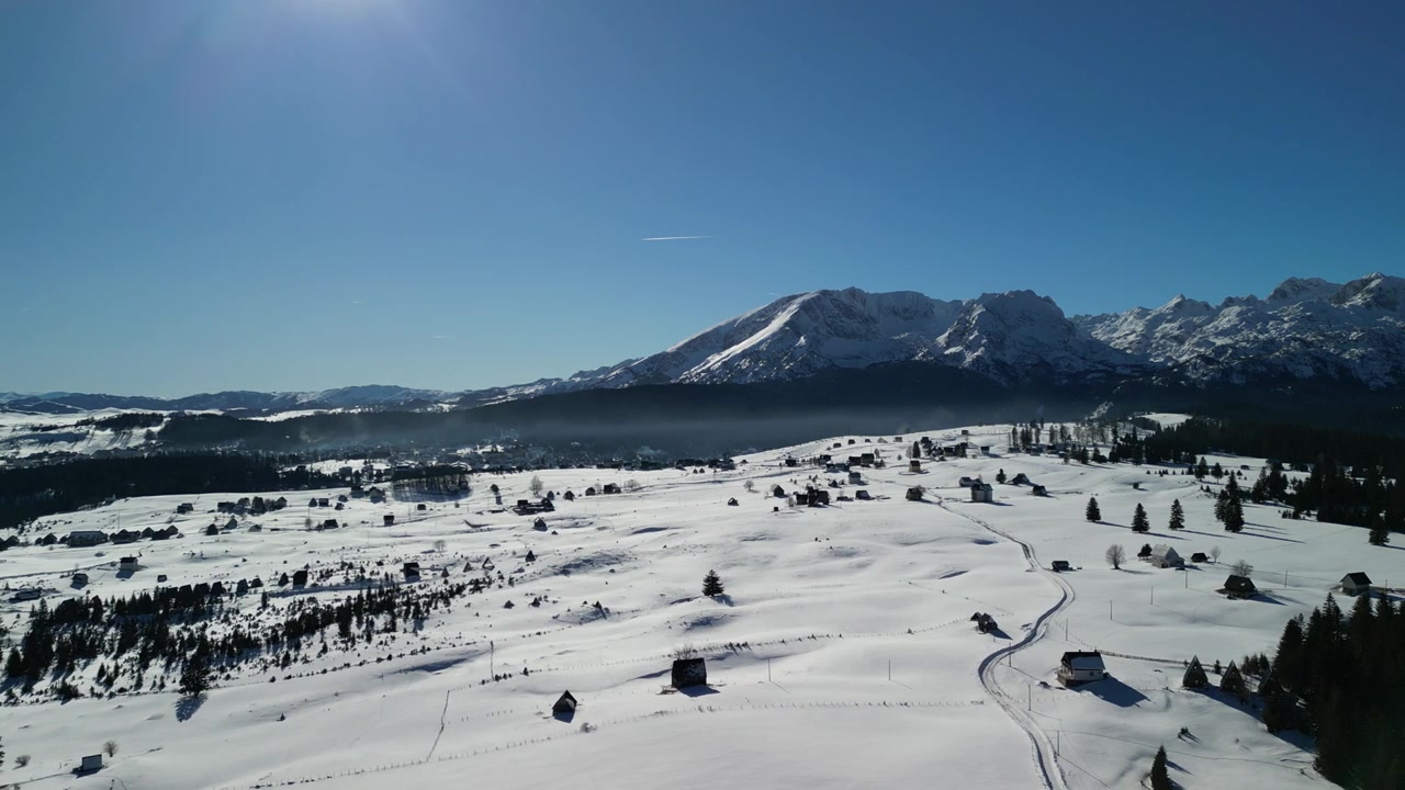 Vista aérea de drone de um vale coberto de neve com cabanas espalhadas, prados abertos e picos de montanhas acidentados sob um céu azul claro no Parque Nacional Durmitor, Montenegro.