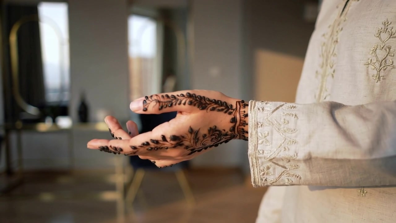 Donna a casa con le mani giunte al petto, che ruota il braccio, mostrando henné e un polsino ricamato, con spazio per il testo.