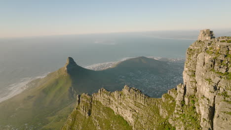 Aerial-footage-of-thin-mountain-ridge-above-city-on-sea-coast.-Rock-wall-lit-by-bright-sunshine.-Cape-Town,-South-Africa