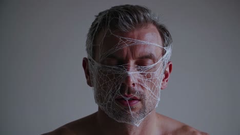 hombre de mediana edad sin camisa con los ojos cerrados y la cara cubierta por una telaraña se mantiene quieto contra un fondo blanco, creando una imagen cautivadora e inquietante
