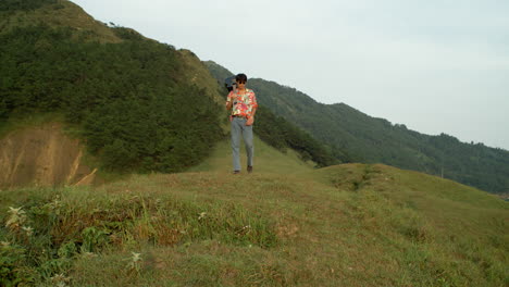 Ein-Junger-Männlicher-Asiatischer-Gitarrenmusiker-Spaziert-Durch-Eine-Natürliche-Grüne-Berglandschaft-Und-Trägt-Sein-Instrument-Entspannt-In-Farbenfroher-Vintage-Kleidung