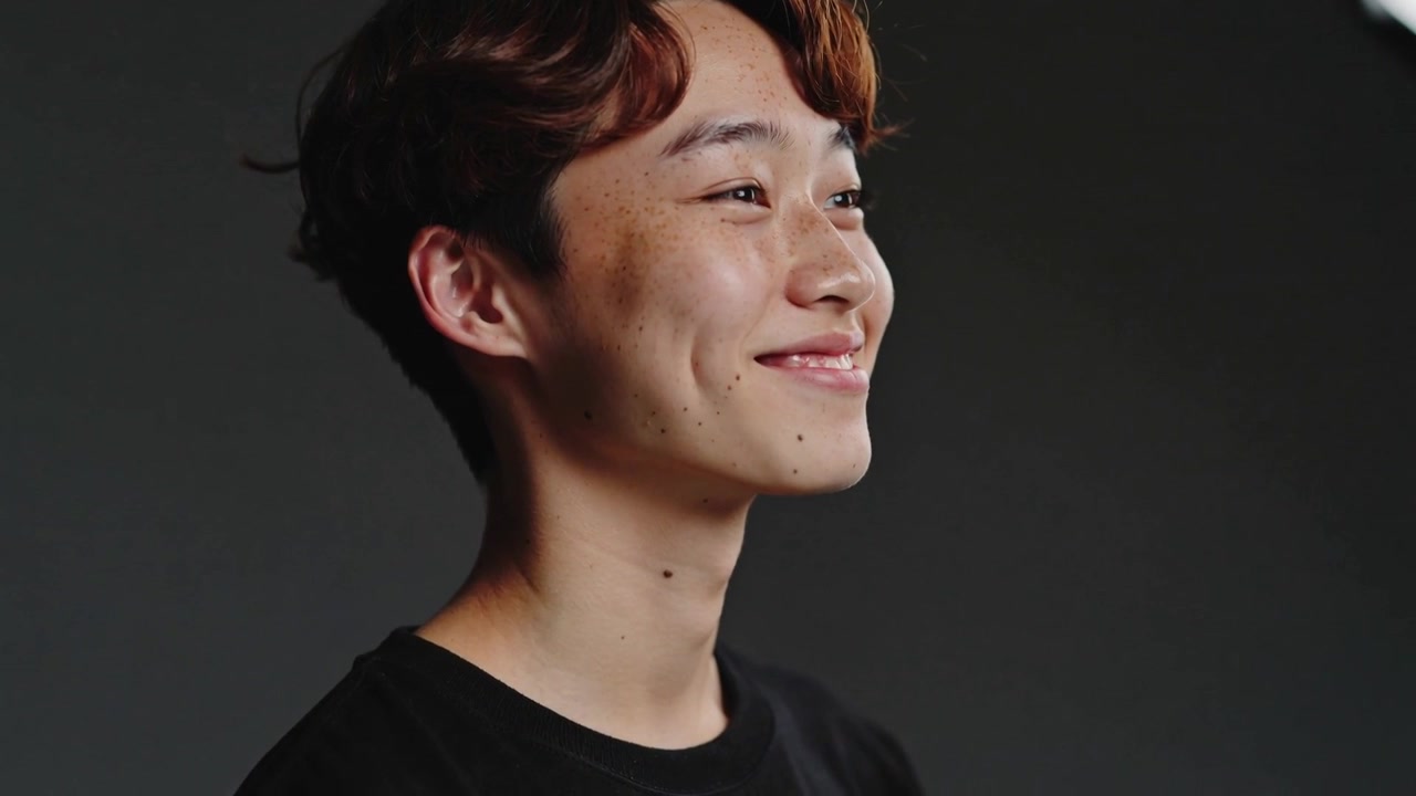 Adolescente asiatico ripreso mentre gira leggermente la testa, sorridente per un ritratto in studio, con softbox, indossando una maglietta nera.