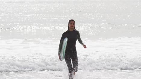 mixed race woman running from the sea onto beach carrying surfboard