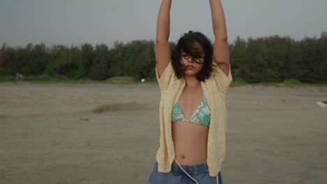 mujer joven estirándose en una playa de arena, árboles en el fondo, día nublado, estado de ánimo sereno
