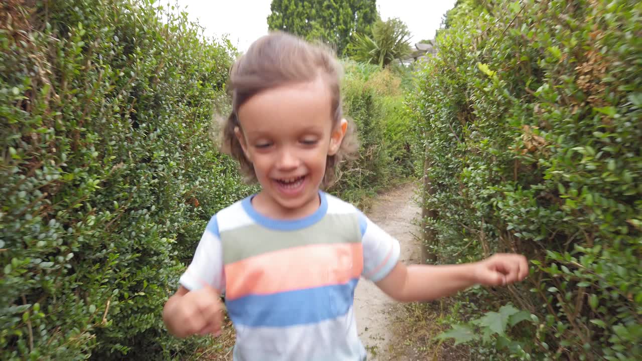 Bebê menino de cabelo comprido correndo em câmera lenta por um labirinto de sebes, vista frontal.