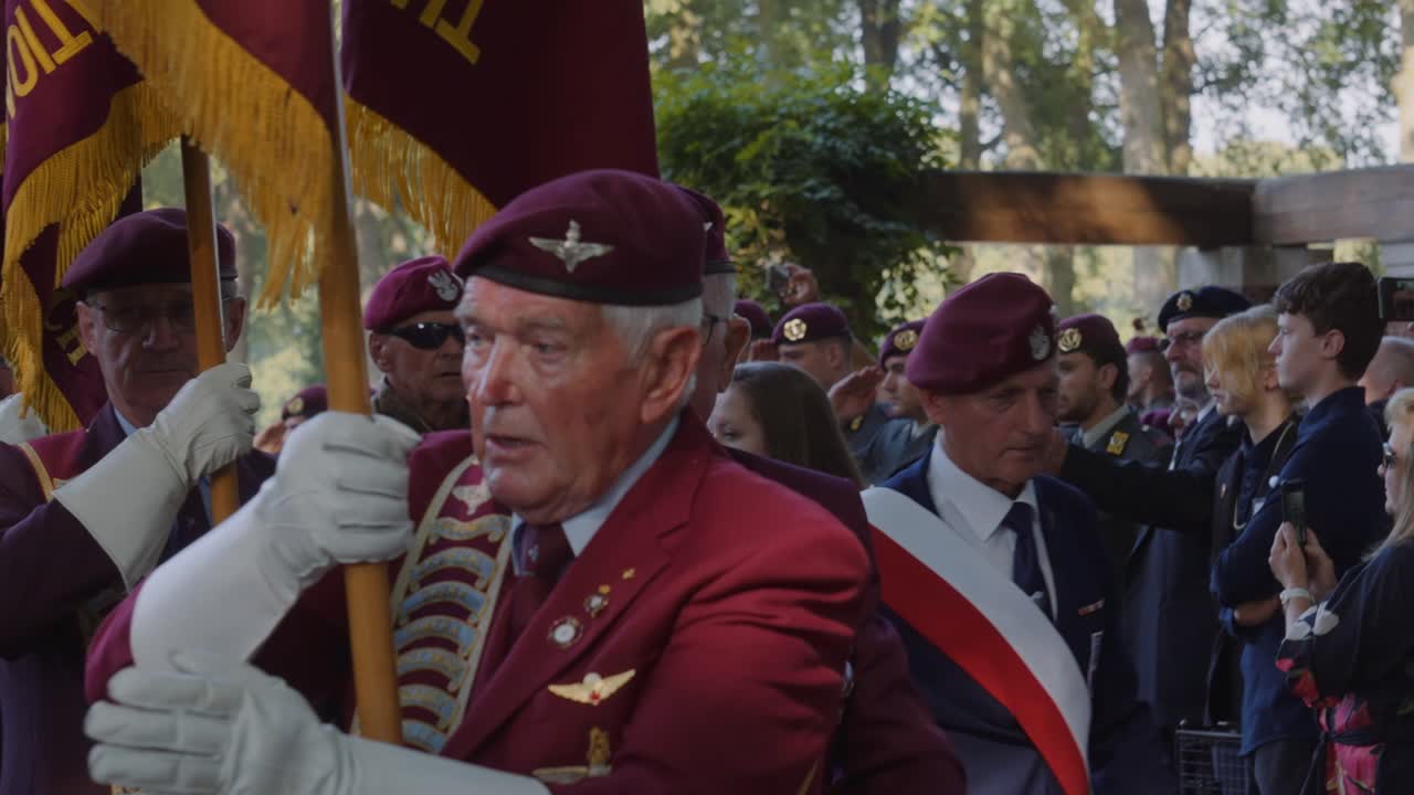 Veteran proudly holds ceremonial flag during memorial service at Oosterbeek War Cemetery. Fellow veterans and military personnel stand in the background, honoring the fallen. Oosterbeek, Gelderland