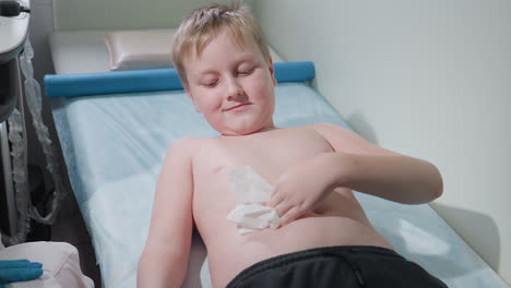 teenage boy wipes chest with tissue after medical examination while seated upright on clinical bed, showing relief and satisfaction following procedure, with medical assistant nearby wearing gloves
