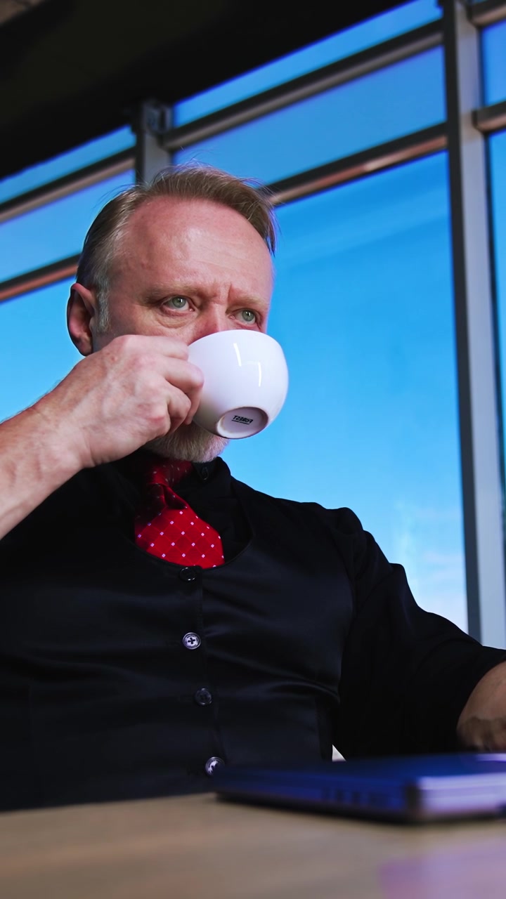 Rolig, selvsikker kaukasisk moden forretningsmand sidder ved skrivebordet på kontoret med en kop i hånden. Manden tager sine briller af, lægger dem på den bærbare computer og drikker kaffe. Lav vinkel. Vertikal video.