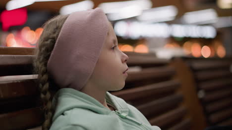 una vista lateral de un niño con una cinta rosa en la cabeza y una sudadera verde, sentado en un banco de madera, mirando atentamente a algo en la distancia, con luz borrosa