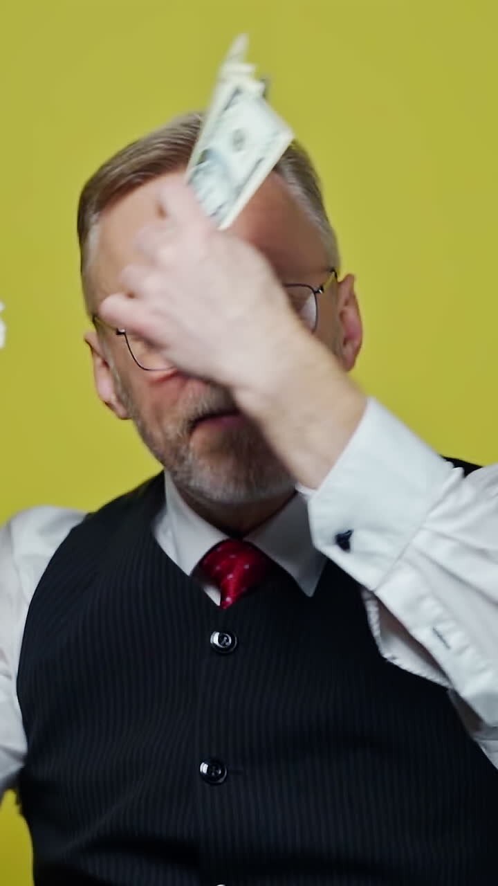 Excited mature man with dollars in hands. Happy elderly man is waving many dollar bills isolated on yellow background. Slow motion. Positive emotions. Vertical video