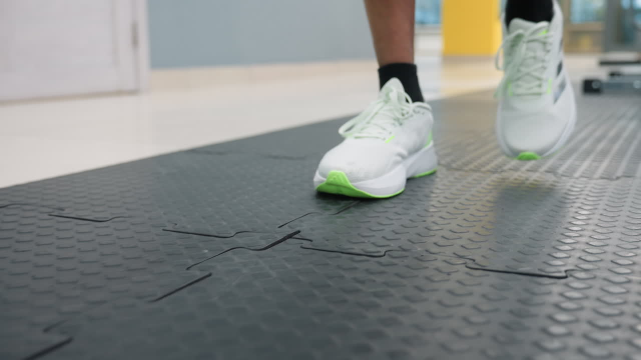 close up of white and neon green sneaker walking on interlocking rubber gym mat next to closed door with wreath, blurred weight machines and bright ceiling lights in background
