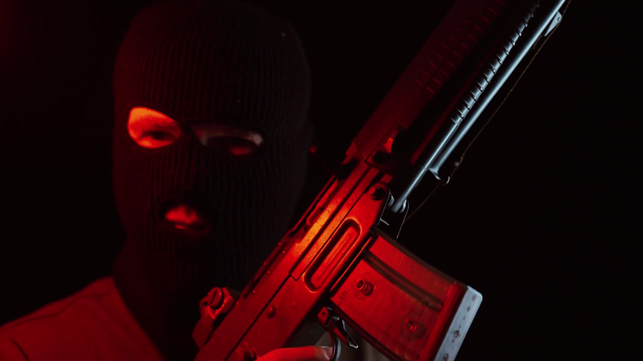 A man wearing a black balaclava holds a rifle illuminated by dramatic red light in a dark setting, creating a cinematic scene filled with mystery, suspense, and intensity