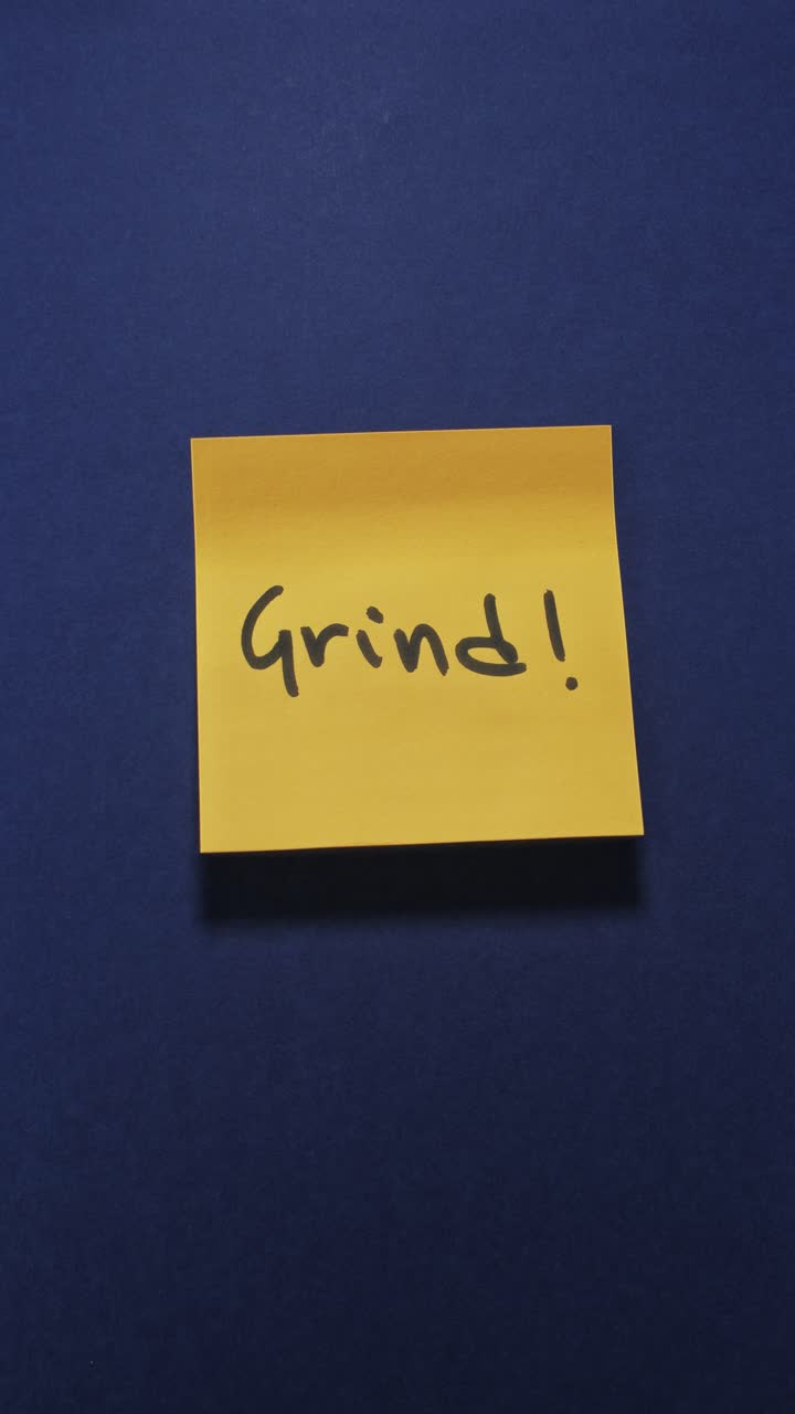 Mãos a colar uma nota adesiva 'Grind' numa parede azul escura, realçando a produtividade.