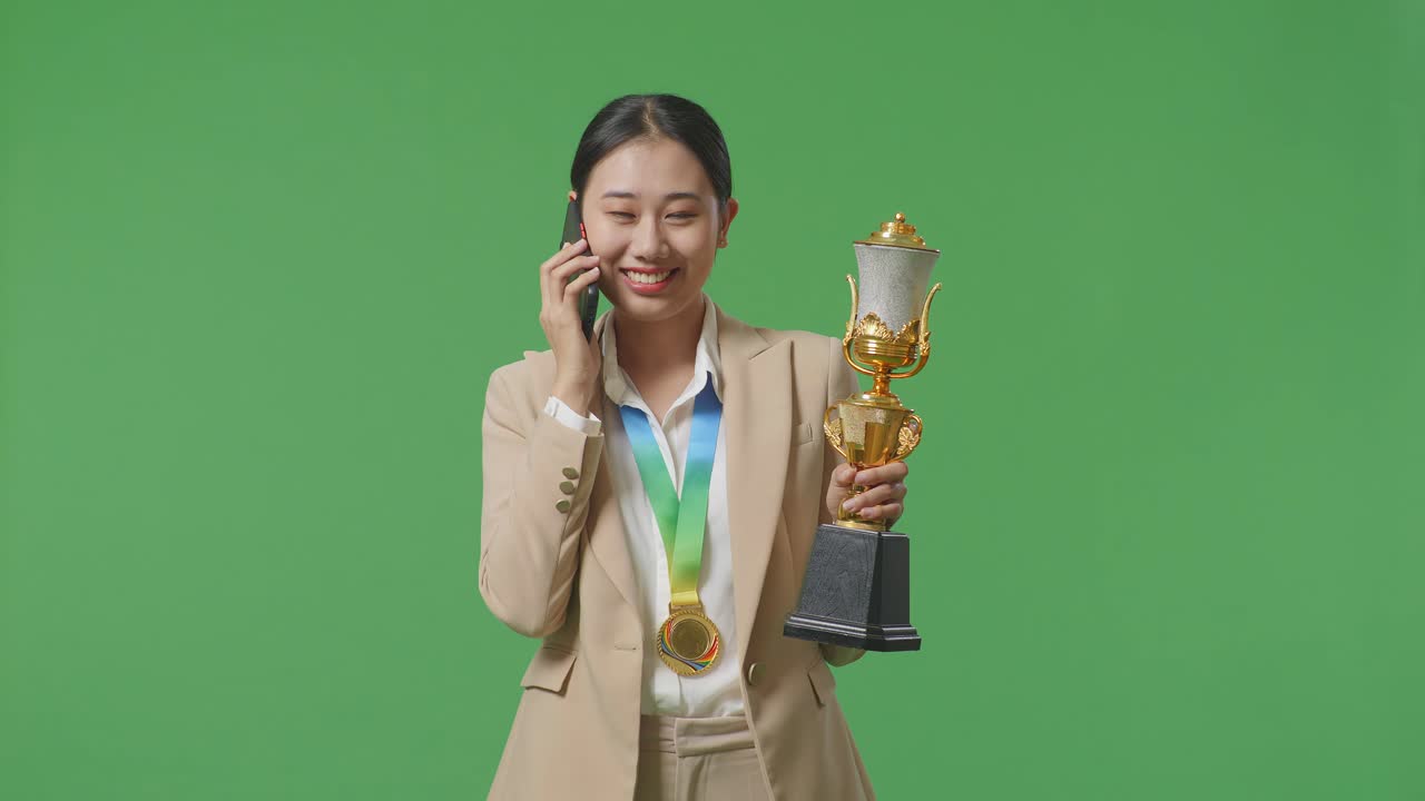 금메달 을 들고 있는 슈트 를 입은 아시아 사업가 여성 이 스튜디오 에서 녹색 스크린 배경 에 있는 스마트폰 에 대화 하고 있다