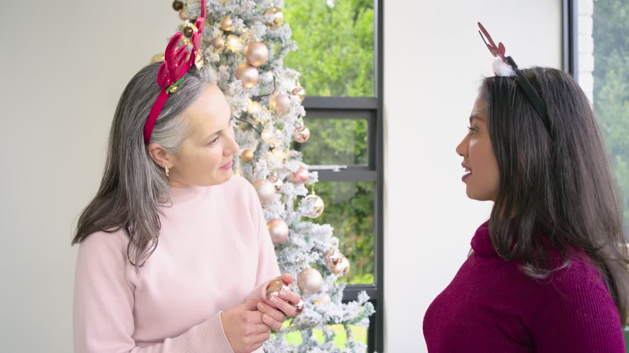 Smiling while exchanging ornaments, diverse female friends decorating tree at home for Christmas