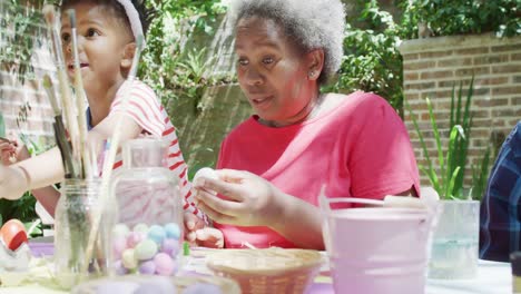 happy african american grandmother and grandson decorating easter eggs in sunny garden, slow motion