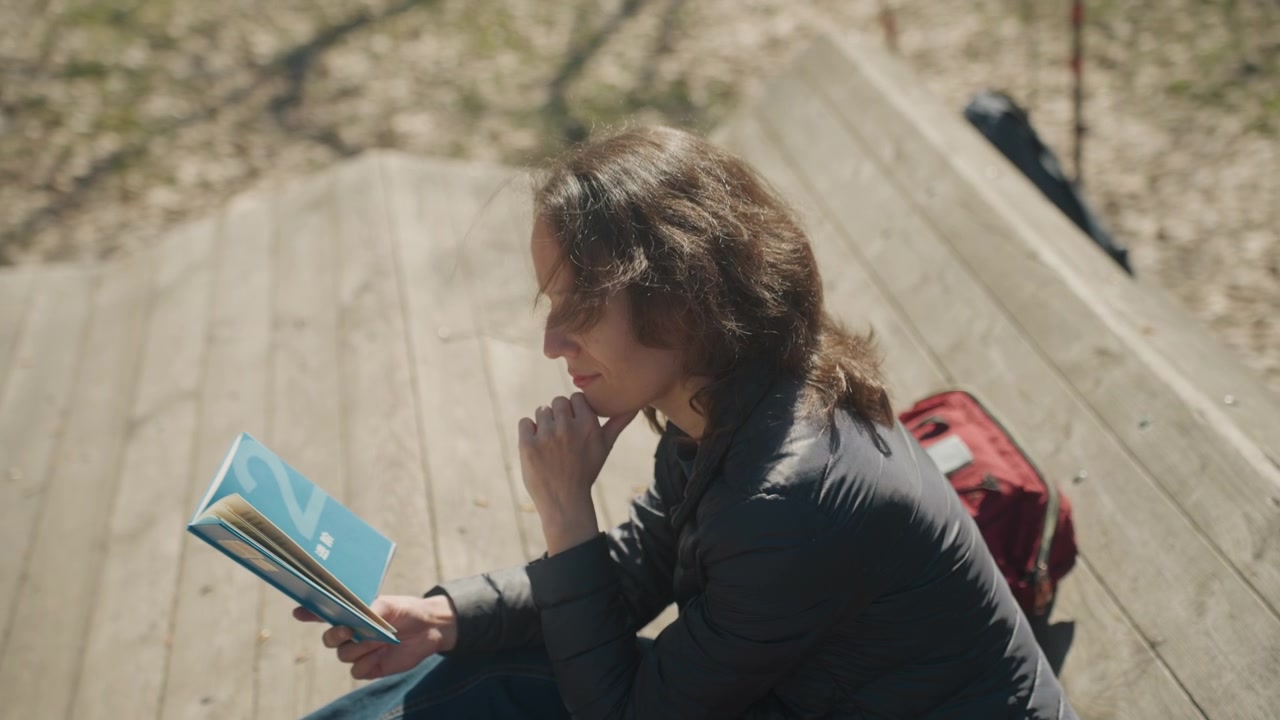 Close up of woman reading book on wooden bench outdoors with red bag nearby. Book has blue cover and visible text subject maintains thoughtful pose throughout. Natural daylight illuminates