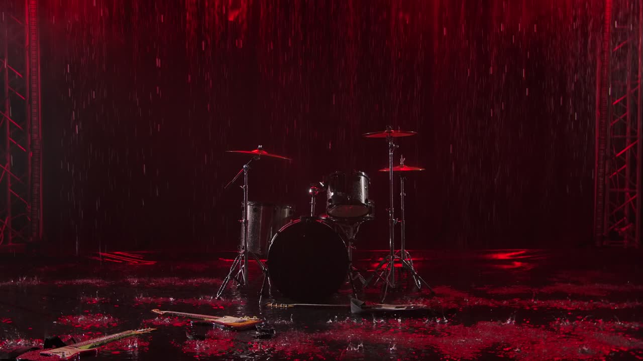 Strumenti musicali abbandonati giacciono sulla superficie dell'acqua sotto la pioggia. Batteria, chitarra elettrica e basso in uno studio buio sono illuminati da una luce rossa. Gocce di pioggia cadono al rallentatore
