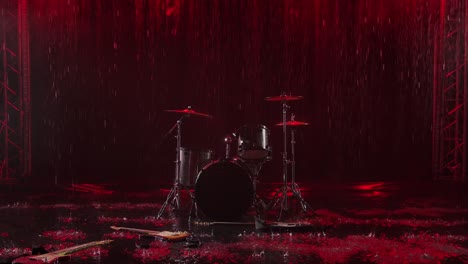 abandoned musical instruments lie on the surface of the water in the rain. drum set, electric and bass guitar in a dark studio are painted with red light. raindrops fall down in slow motion