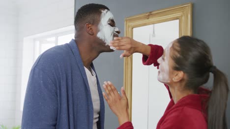 video de una feliz pareja diversa divirtiéndose aplicando máscaras faciales de limpieza y riendo en el baño