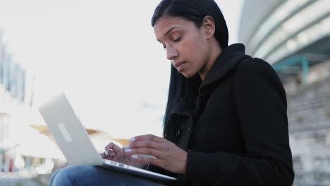 Señora-Hindú-Enfocada-Escribiendo-En-Una-Computadora-Portátil-Mientras-Está-Sentada-Al-Aire-Libre