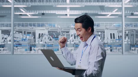 side view of an asian male professional worker standing with his laptop at factory digitalization, he raises his fist up with screaming goal after typed on the laptop