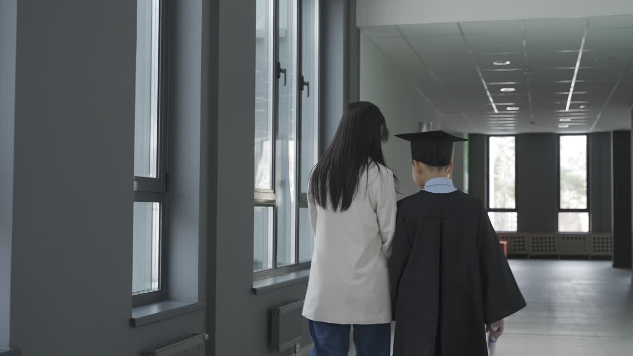 Asian mom hugging and walking with her son. He's wearing a gown and a mortarboard.