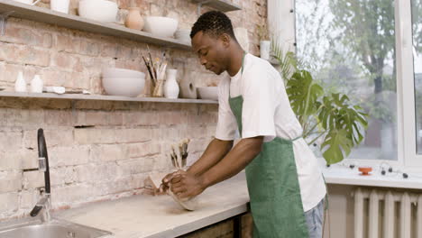 empleado americano en un delantal verde amasando arcilla encima de una mesa en un taller de cerámica